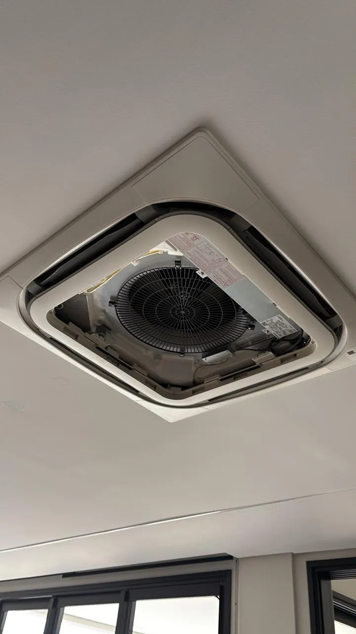 Ar-condicionado cassete no teto, com a tampa aberta e os componentes internos visíveis.