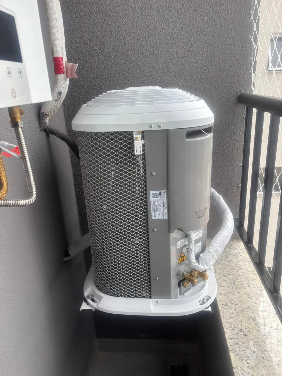 Condensadora instalada em uma varanda, com tubulação isolada e conexões visíveis.